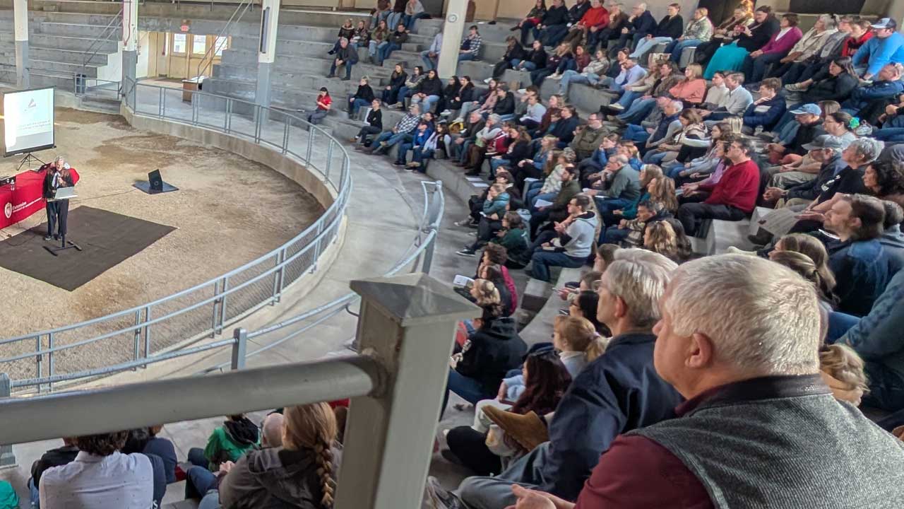 A large crowd, seated in a semi-circular livestock pavilion, is shown watching Dr. Temple Grandin speak.